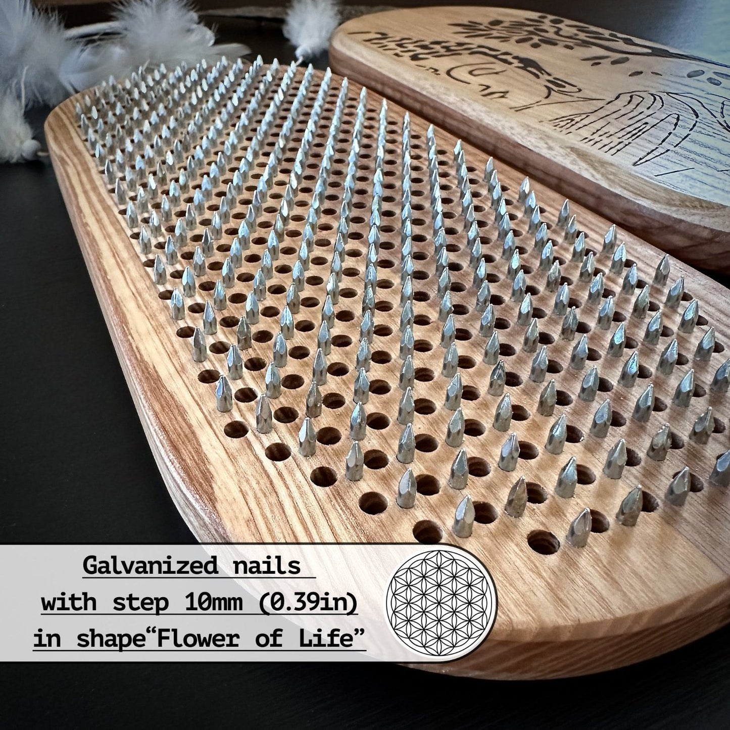 Close-up view of a Sadhu board "Buddha" featuring galvanized nails with a 10mm (0.39in) step, arranged in the shape of the "Flower of Life." The board is made from natural ash wood, with an engraved Buddha and tree design on the other side. White feathers and a dream catcher are visible in the background, enhancing the spiritual theme