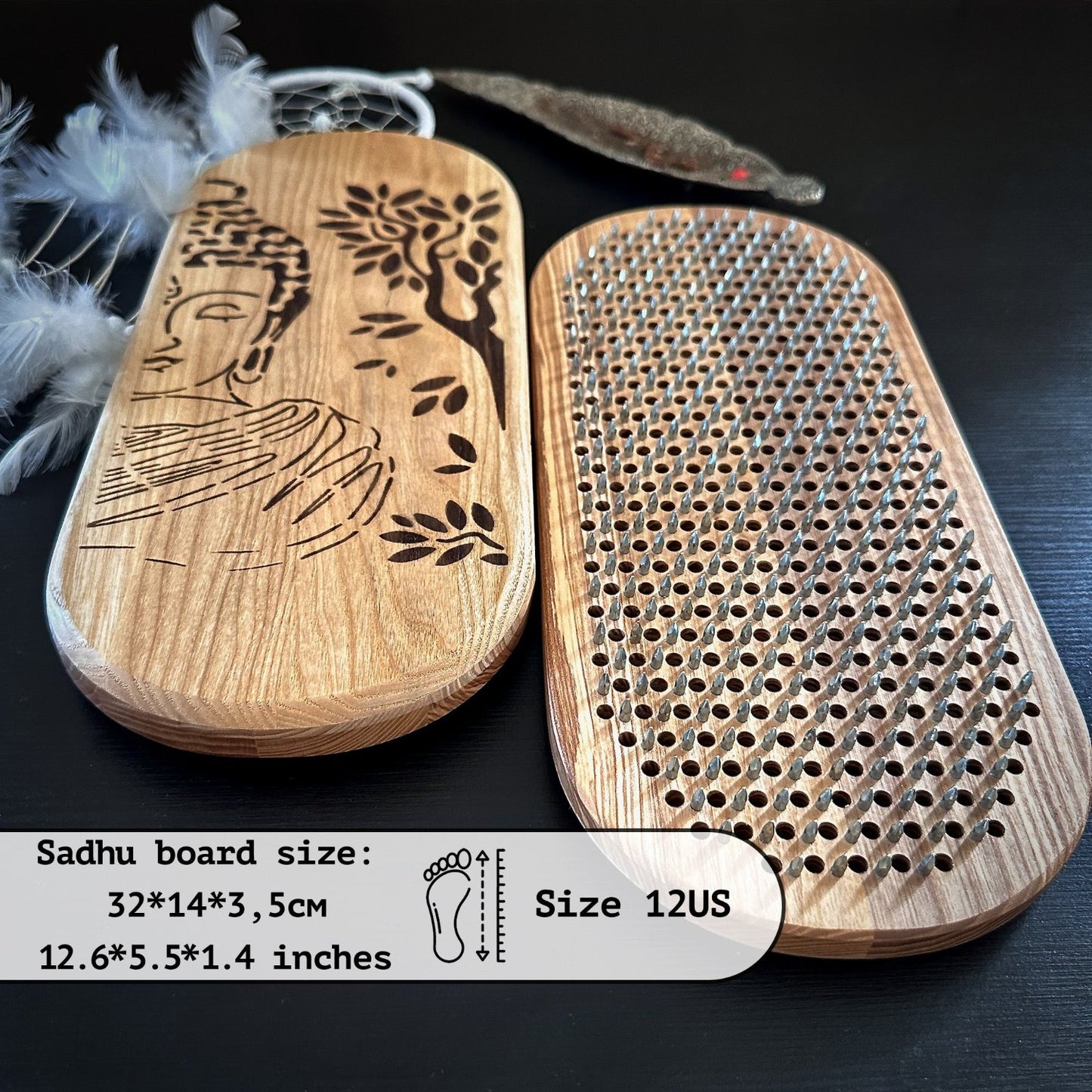 a pair of Sadhu boards, which are wooden boards used for standing on nails. The board on the left features an engraved design of a Buddha under a tree, while the board on the right has a grid of evenly spaced metal nails. The dimensions of the Sadhu boards are specified as 32 cm by 14 cm by 3.5 cm (12.6 inches by 5.5 inches by 1.4 inches), and they are designed to accommodate a foot size of 12 US. The background includes a feathered dreamcatcher and another decorative element.