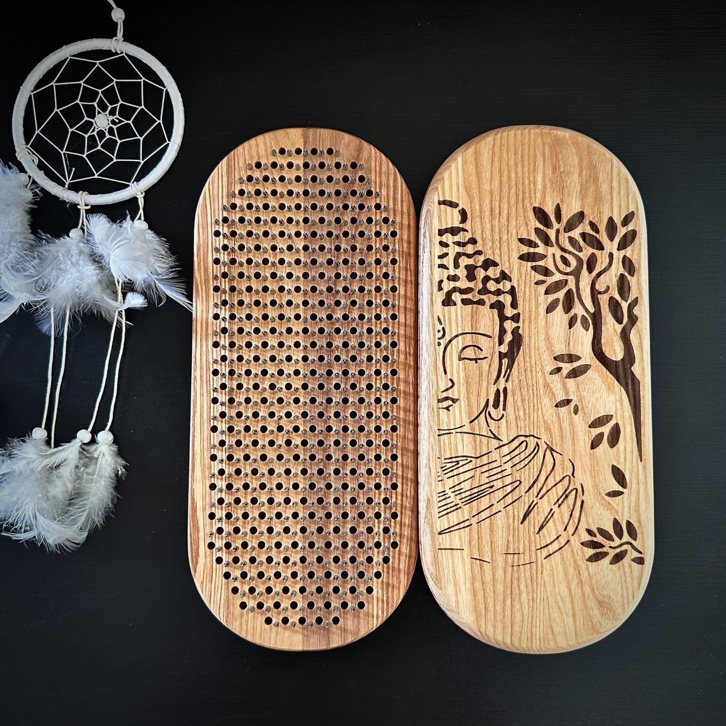 The image shows a set of Sadhu boards, one with an engraved Buddha design and the other with metal nails. A feathered dreamcatcher is visible in the background.