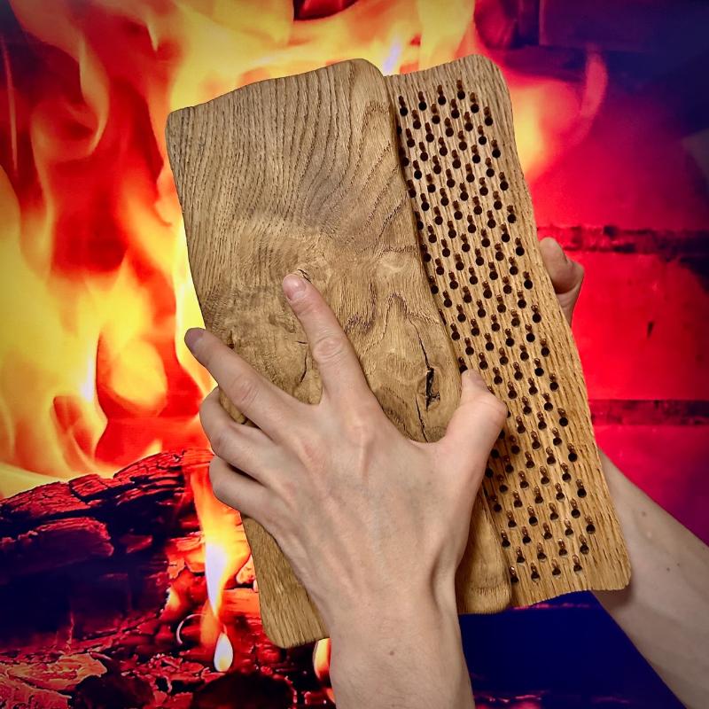 A pair of hands holding two rustic wooden sadhu boards, one of which features rows of small holes for copper nails, with a fiery background creating a dramatic and intense atmosphere.