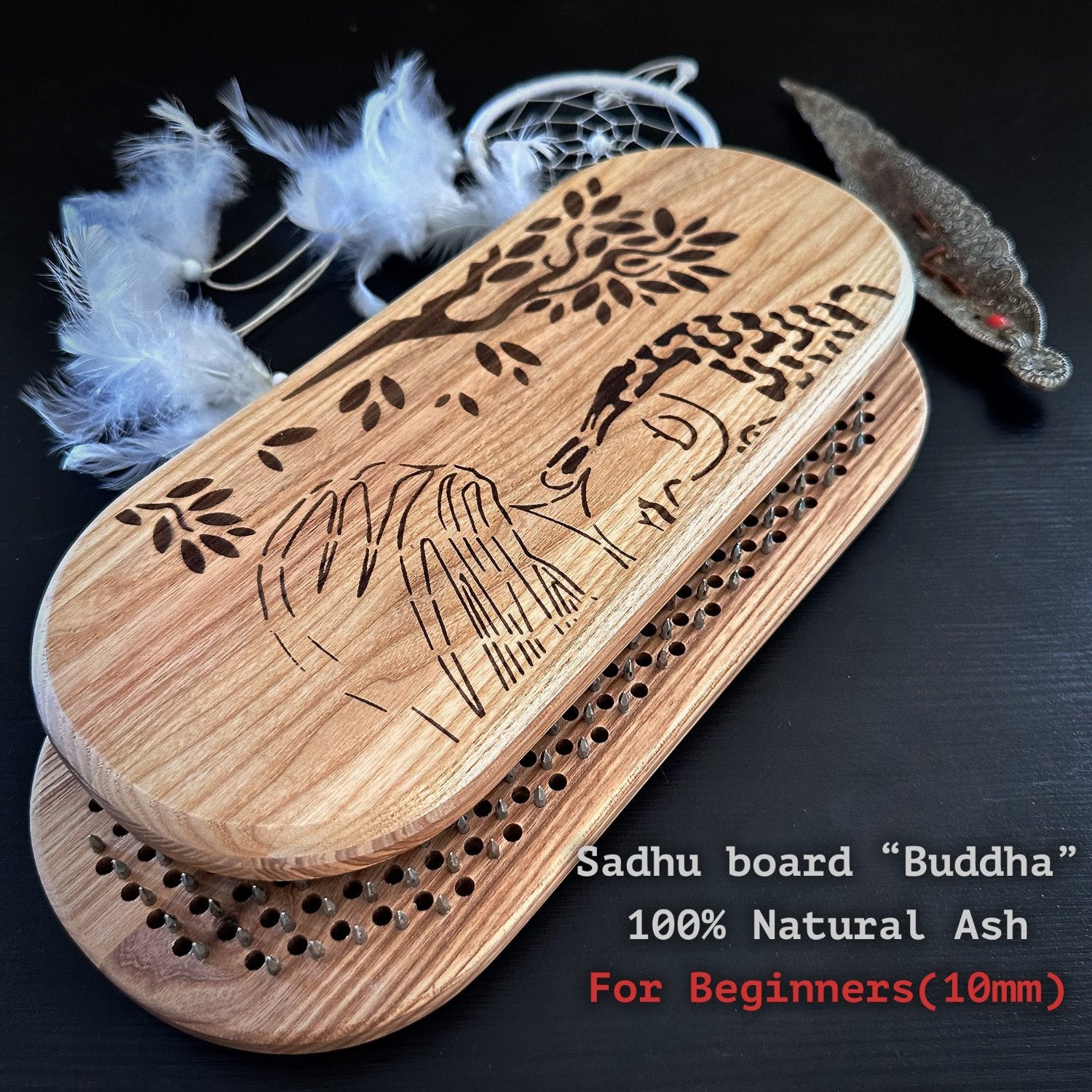 Buddha" made from 100% natural ash wood, featuring an engraved design of Buddha and a tree. The board is designed for beginners with a 10mm spacing between the holes for nails. The setup includes a dream catcher with white feathers and an incense holder, adding to the spiritual ambiance