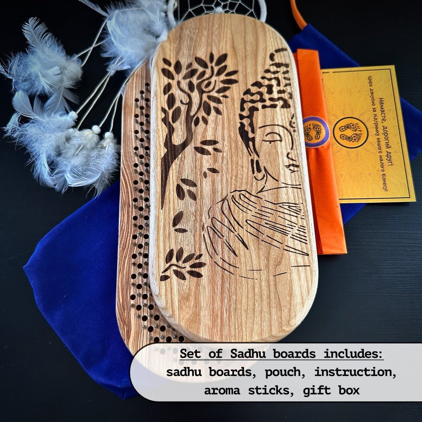 The image shows a set of Sadhu boards, one with an engraved Buddha design and the other with metal nails. The set includes a blue pouch, instructions, aroma sticks, and a gift box. A feathered dreamcatcher is visible in the background. The text at the bottom states the contents of the set: Sadhu boards, pouch, instructions, aroma sticks, and a gift box.