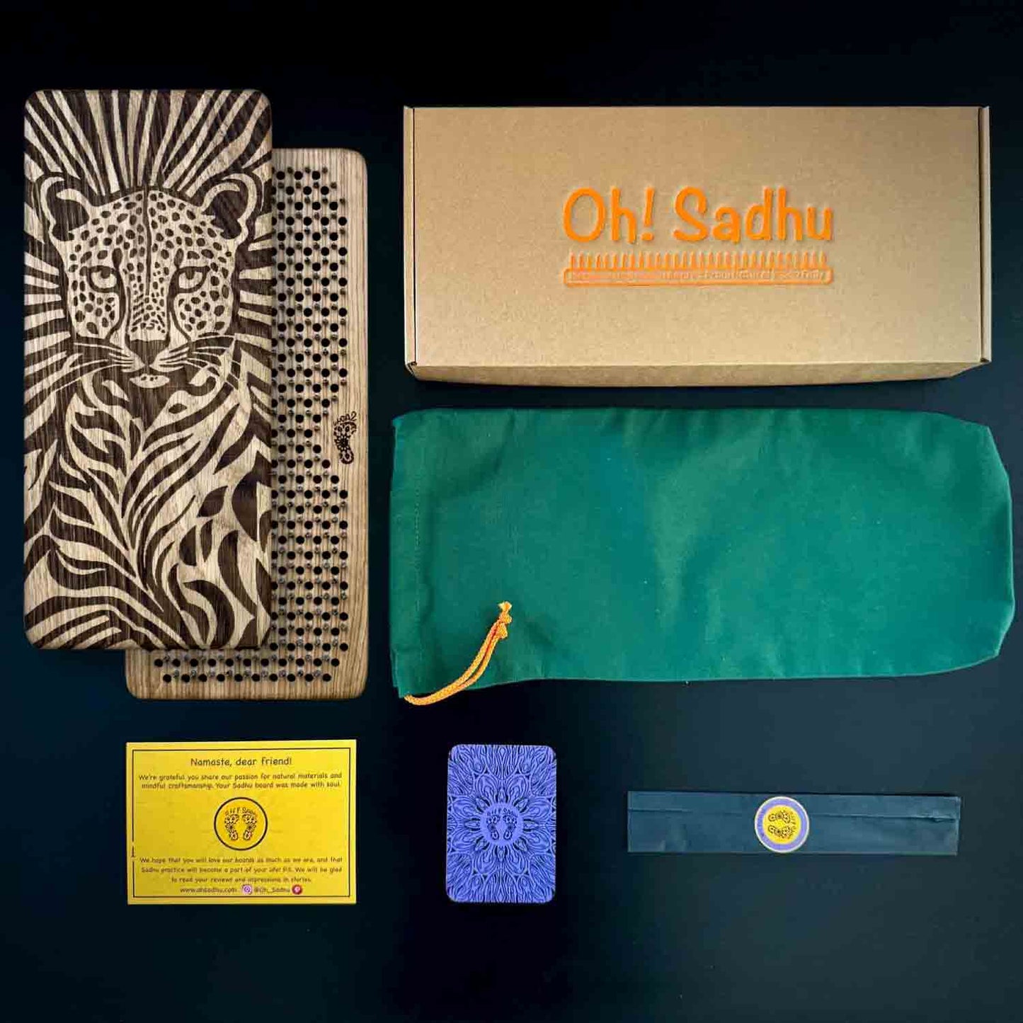 A flat lay of a yoga gift set on a dark background, including a laser-engraved Sadhu boards with cheetah, a kraft paper box labeled 'Oh! Sadhu', a green drawstring pouch, a yellow instruction card, a blue metaphorical card, and incense sticks in black paper wrap.