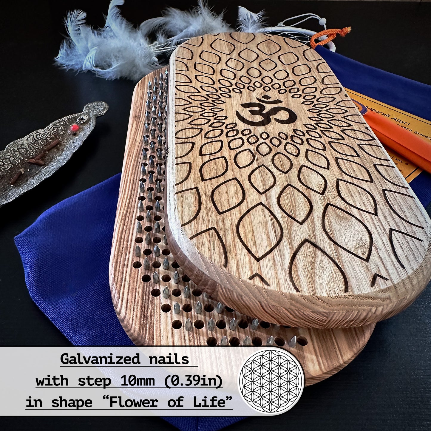 A set of Sadhu boards with an engraved Om symbol and galvanized nails arranged in the "Flower of Life" pattern, with a 10mm (0.39in) step. The set includes a pair of boards, a blue pouch, aroma sticks, an instruction card, and a decorative gift box. Feathers and a metal incense holder are also shown in the background.