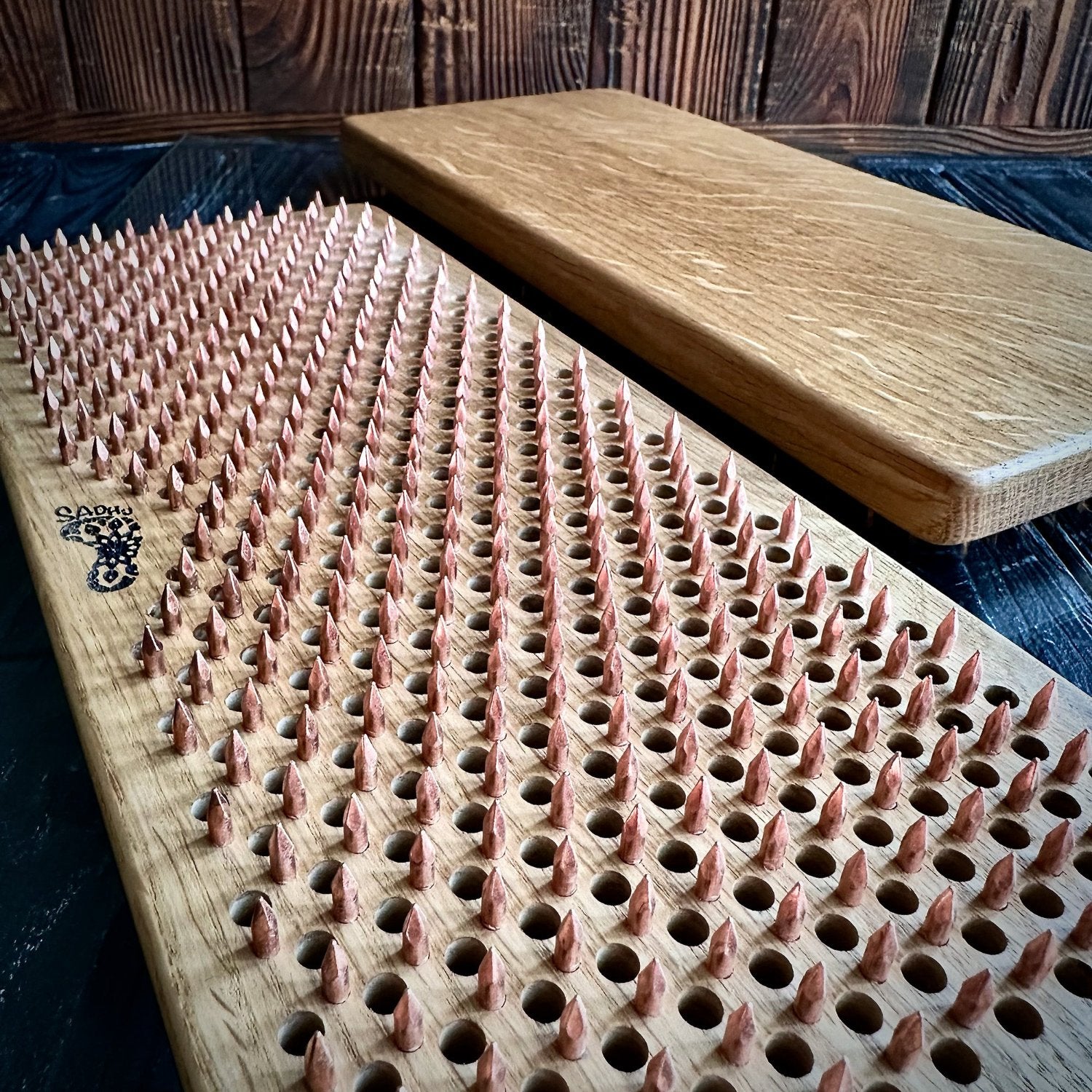 Two Sadhu Boards made from natural oak wood, featuring a smooth finish and copper nails. One board is shown with a close-up view of the nail pattern, while the other displays the natural grain and texture of the oak. Sadhu boards are set against a dark, rustic wooden background.