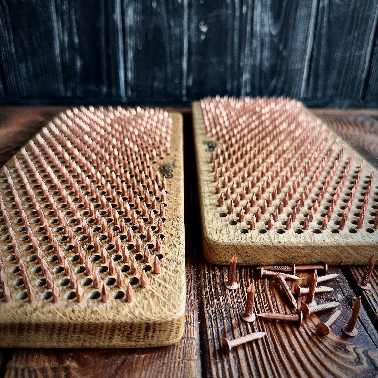 Close-up view of two Sadhu Boards placed side by side, each featuring rows of evenly spaced copper nails embedded into natural oak wood. The boards display intricate nail patterns designed for acupressure, set against a rustic, dark wooden background. A few loose copper nails are scattered in the foreground, highlighting the craftsmanship and unique details of the boards.