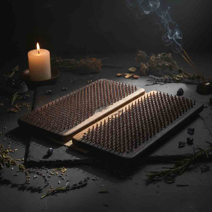 Two wooden sadhu nail boards styled on a dark surface with a lit candle, dried botanicals, and a smoking incense stick in yoga studio