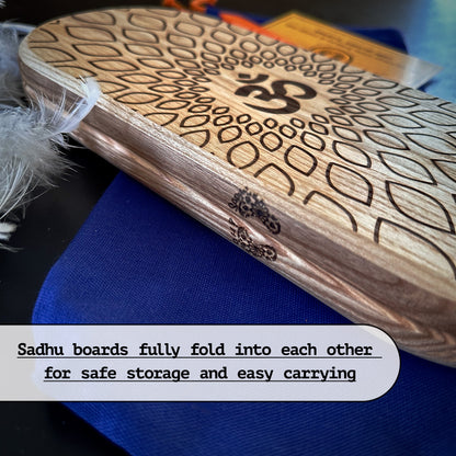 The image shows a pair of Sadhu boards with an Om symbol and lotus design, folded together for safe storage and easy carrying. The boards are placed on a blue pouch, with a feathered dreamcatcher and some booklets in the background. The text at the bottom highlights the boards' ability to fold into each other.