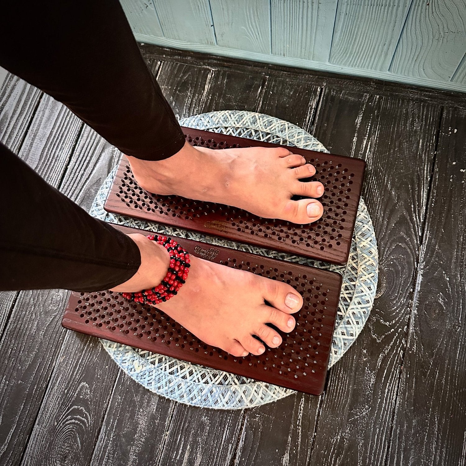 A person standing barefoot on two Sadhu boards with a grid of nails, with one foot wearing a red beaded anklet. Sadhu boards are placed on a round patterned mat on a dark wooden floor.