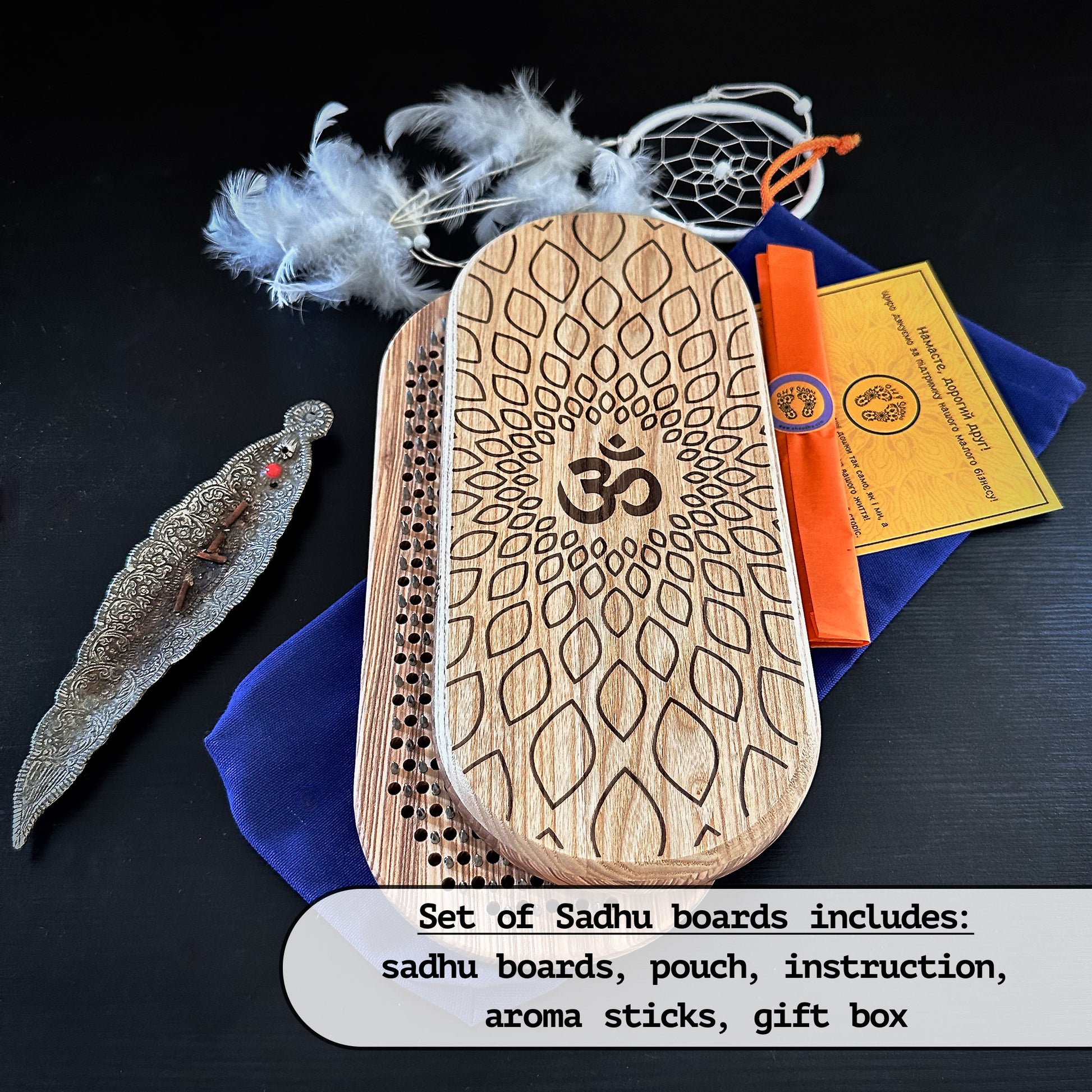 A set of Sadhu boards made from natural ash wood, featuring an engraved Om symbol. Includes a pair of boards, a blue pouch, aroma sticks, an instruction card, and a decorative gift box. Feathers and a metal incense holder are also shown in the background.