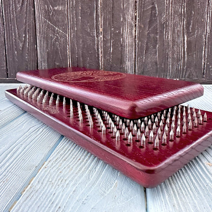 A burgundy-colored Sadhu board with metal nails arranged in a grid pattern, partially open to show the sharp spikes inside. The board features a Tree of Life engraving on the top surface, placed on a light blue wooden floor against a dark wooden wall.