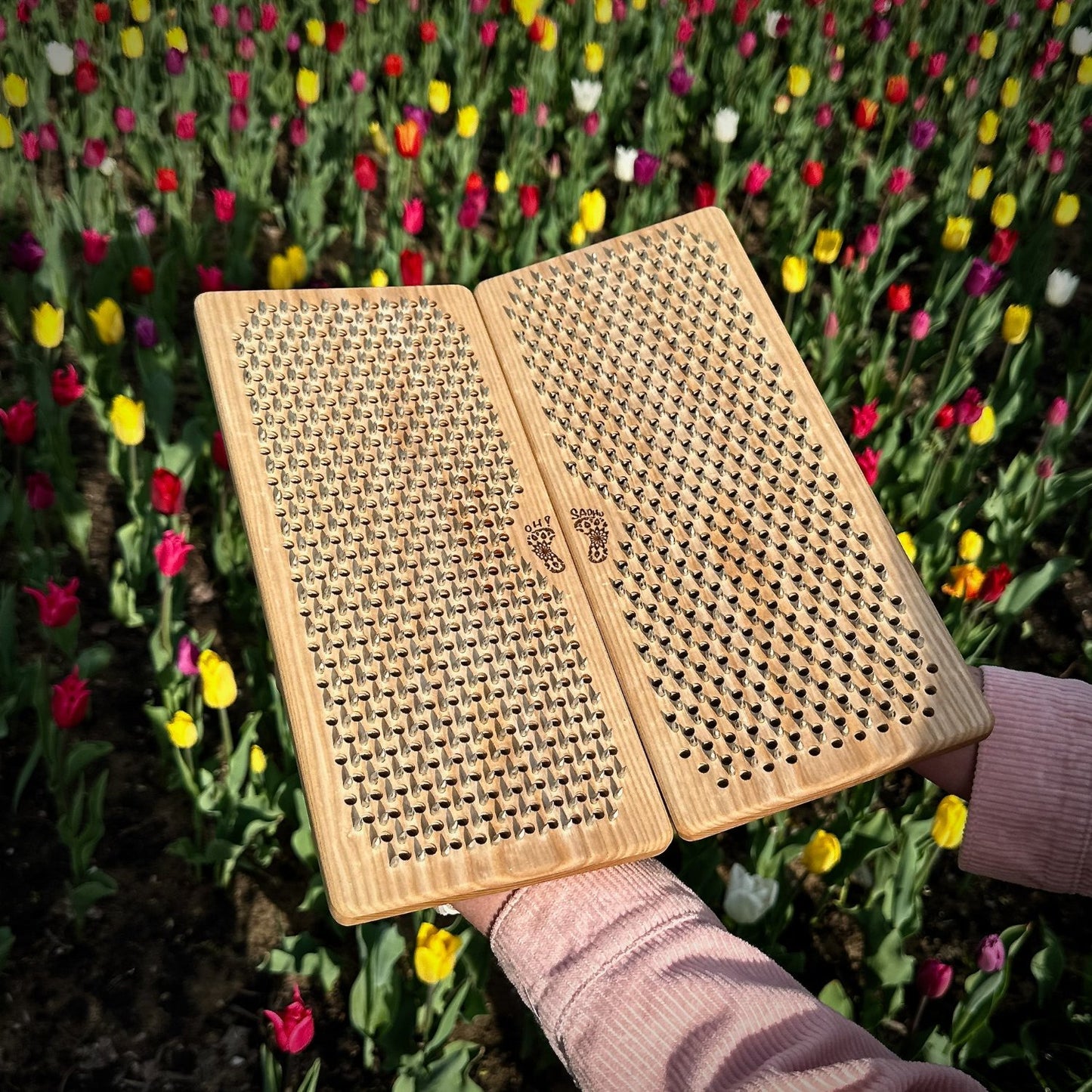 open sadhu board with nails in shape flower of life in hands, with small feet logo engraving on flowerbed of tulip in background