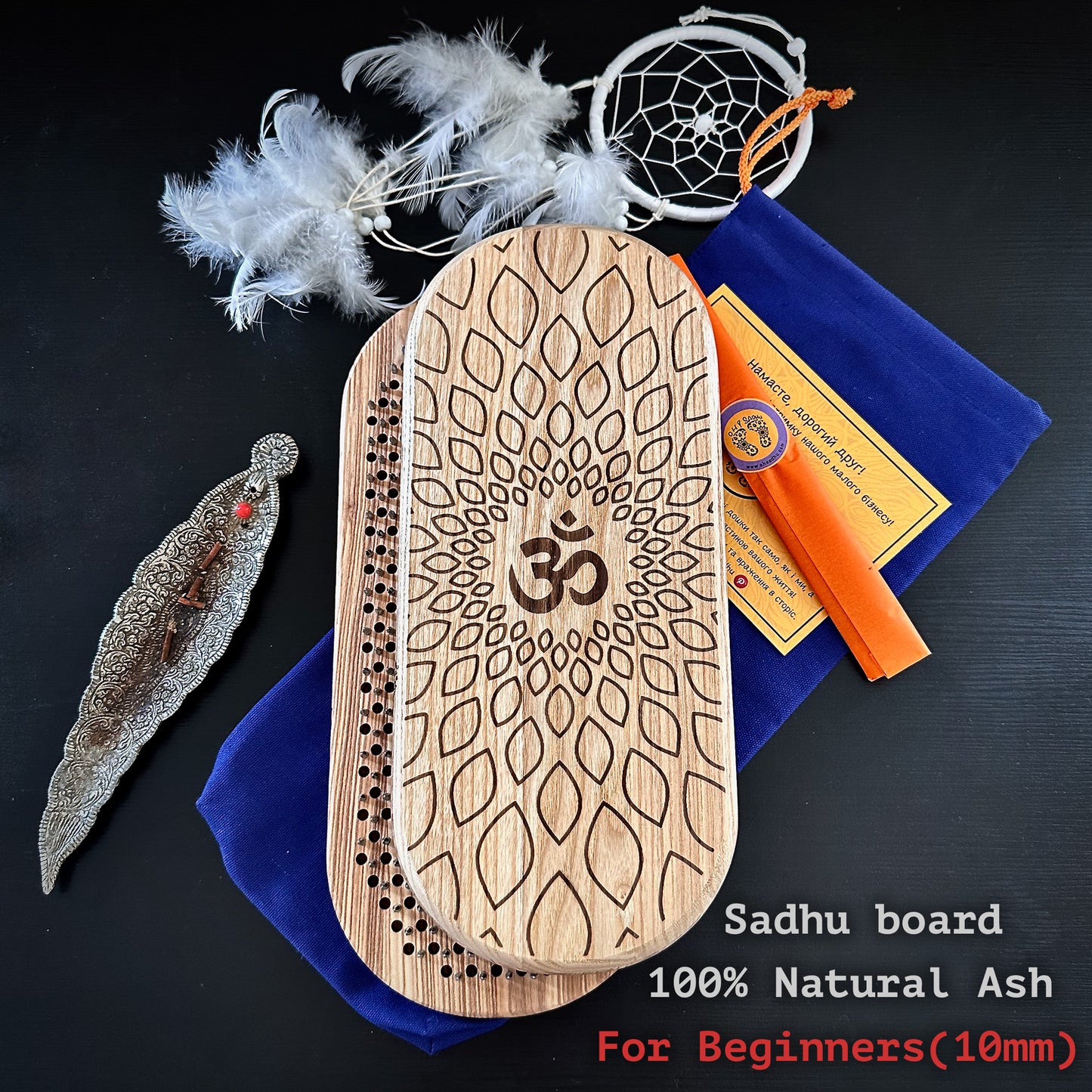 set of Sadhu boards made from 100% natural ash wood, designed for beginners with 10mm nail spacing. One board features an Om symbol and lotus design, while the other has evenly spaced nails. The set od sadhu boards includes a blue pouch, instructions, aroma sticks, and a feathered dreamcatcher in the background. The text highlights that sadhu boards are suitable for beginners.